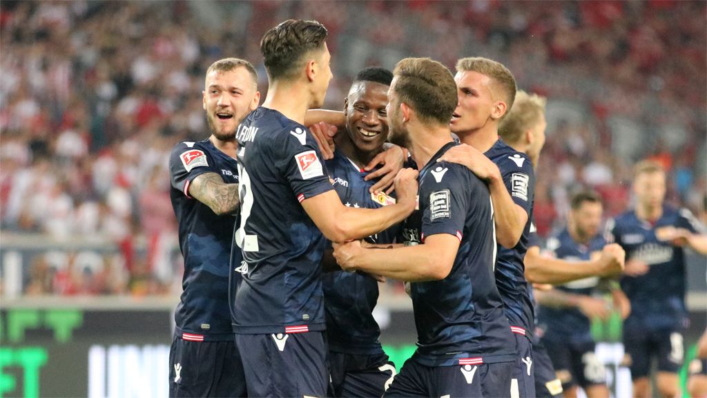Spieler einer Fußballmannschaft feiern gemeinsam ein Tor auf dem Spielfeld, umgeben von jubelnden Fans.