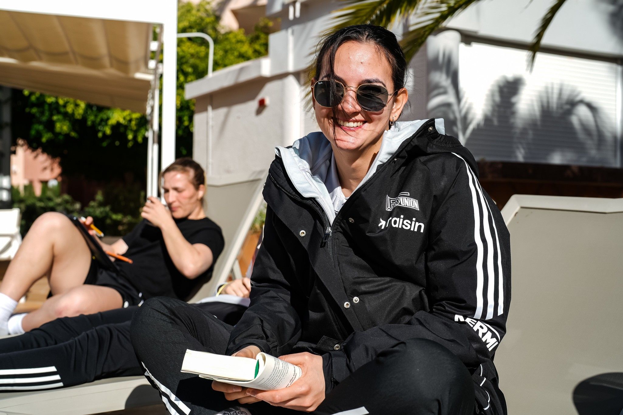 Eine Person mit Sonnenbrille und einem schwarzen Mantel lächelt, während sie ein Buch hält. Im Hintergrund sitzt eine weitere Person.