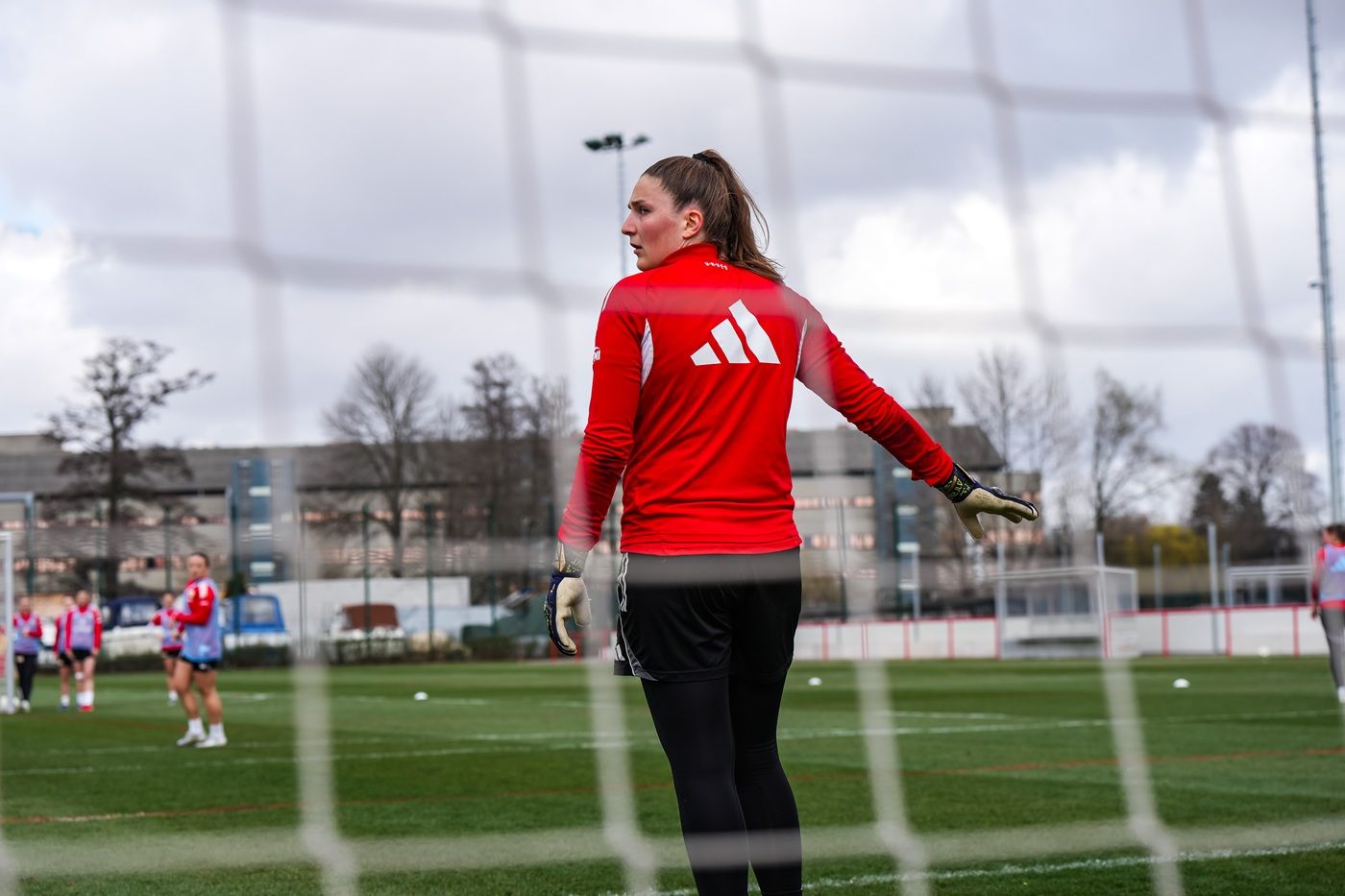 Eine Torhüterin in rotem Trikot steht im Tor während eines Fußballtrainings auf einem Rasenplatz.