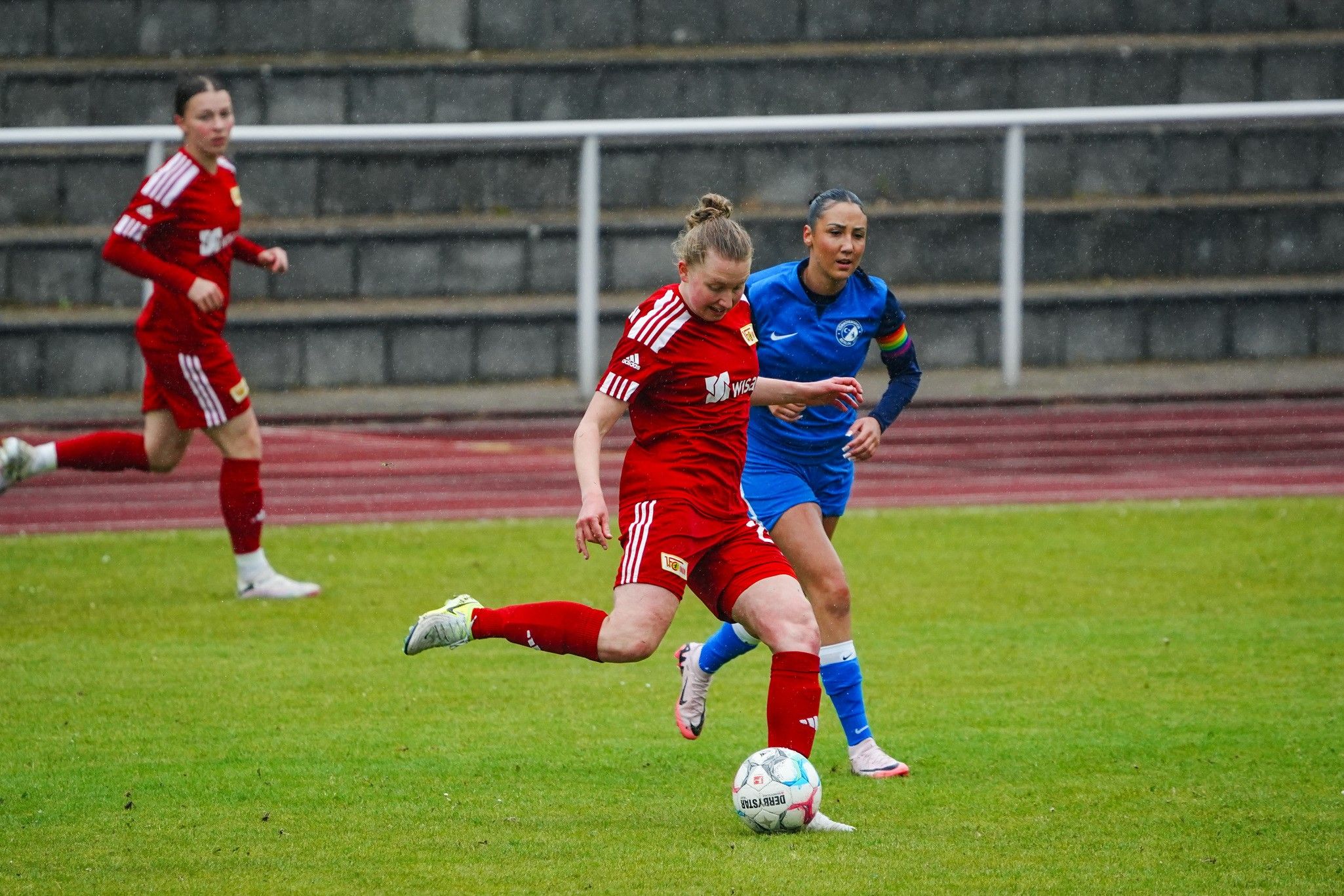 Spielerinnen in Rot und Blau stehen auf einem Fußballfeld. Eine Spielerin in Rot tritt den Ball. Es regnet.