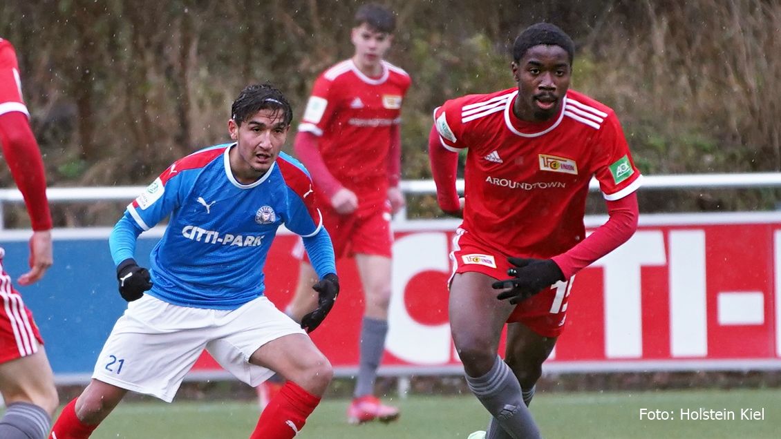 Zwei Fußballspieler in roten und blauen Trikots laufen auf dem Platz; im Hintergrund ist ein weiterer Spieler sichtbar.