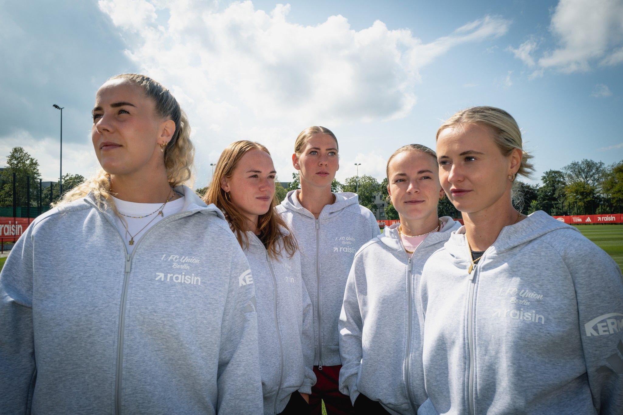 Fünf sportliche Frauen in grauen Hoodies stehen zusammen auf einem Fußballplatz unter einem bewölkten Himmel.