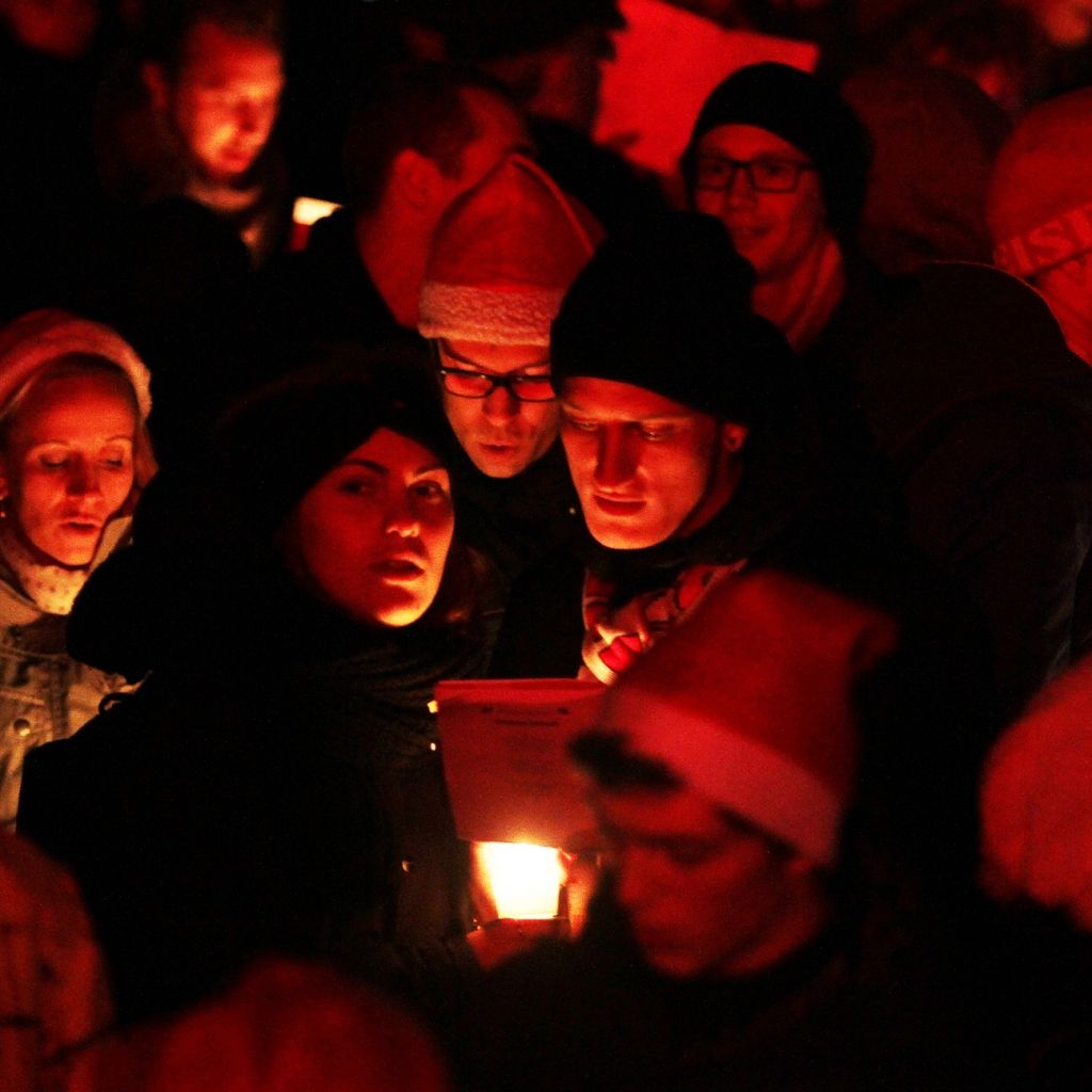 Eine Gruppe von Menschen steht dicht beieinander, einige halten Kerzen. Die Szenerie ist in rotes Licht getaucht.