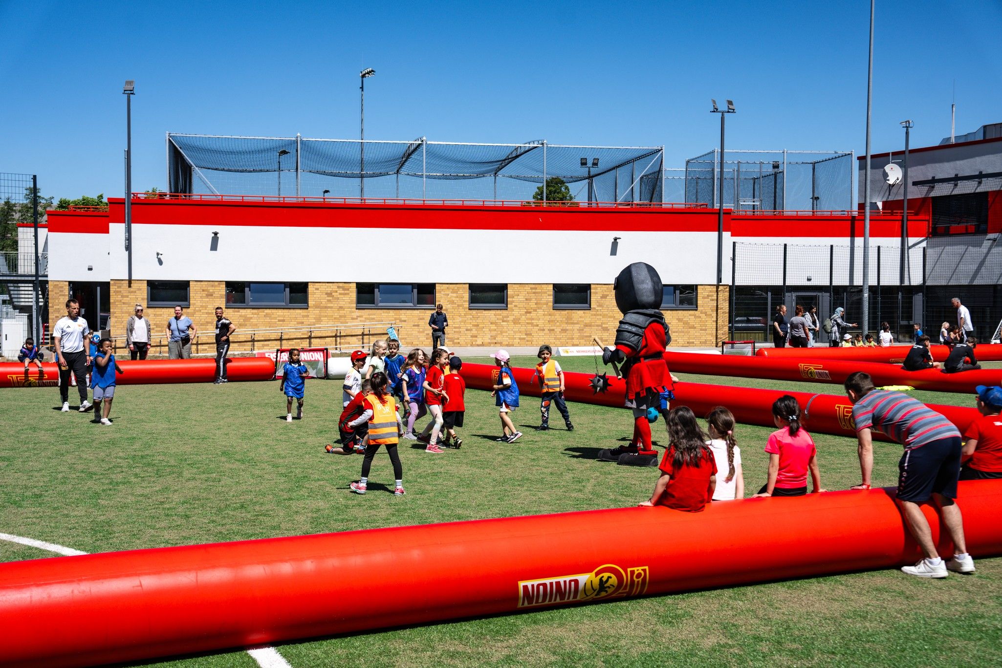Kinder spielen auf einem Sportplatz mit roten Banden, während ein Maskottchen in der Mitte steht. Zuschauer beobachten das Geschehen.