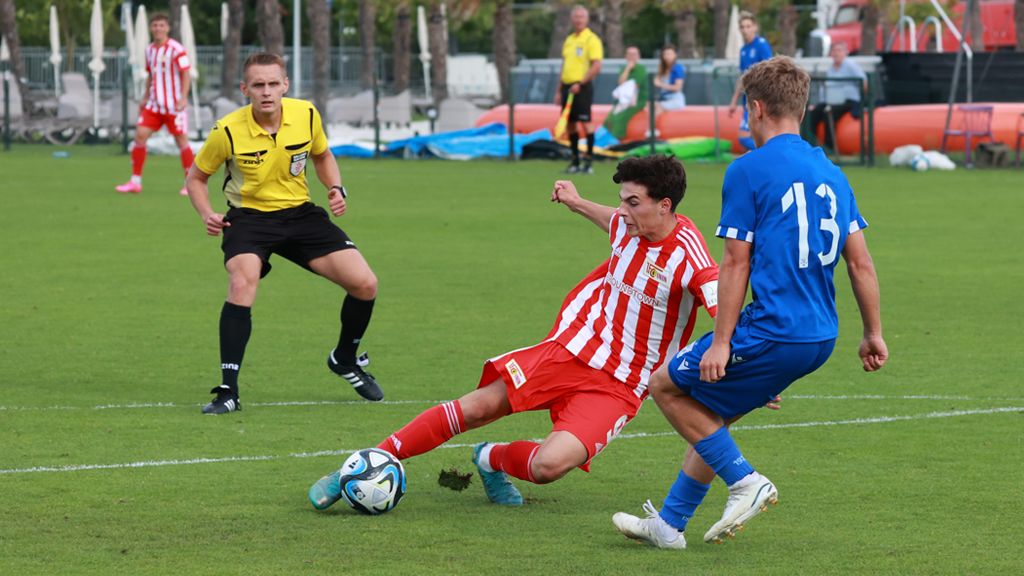 Fußballspiel mit einem Spieler in roten Streifen, der rutscht, während ein blauer Spieler ihn verteidigt. Ein Schiedsrichter beobachtet die Szene.