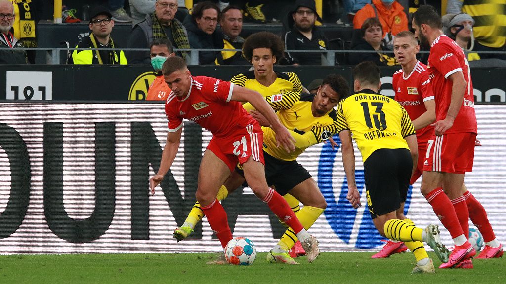 A soccer match between two teams, with players in yellow and red jerseys chasing a ball in midfield.