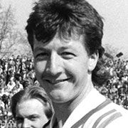 Ein lächelnder Mann mit kurzen, welligen Haaren, posed vor einer Menschenmenge im Hintergrund.