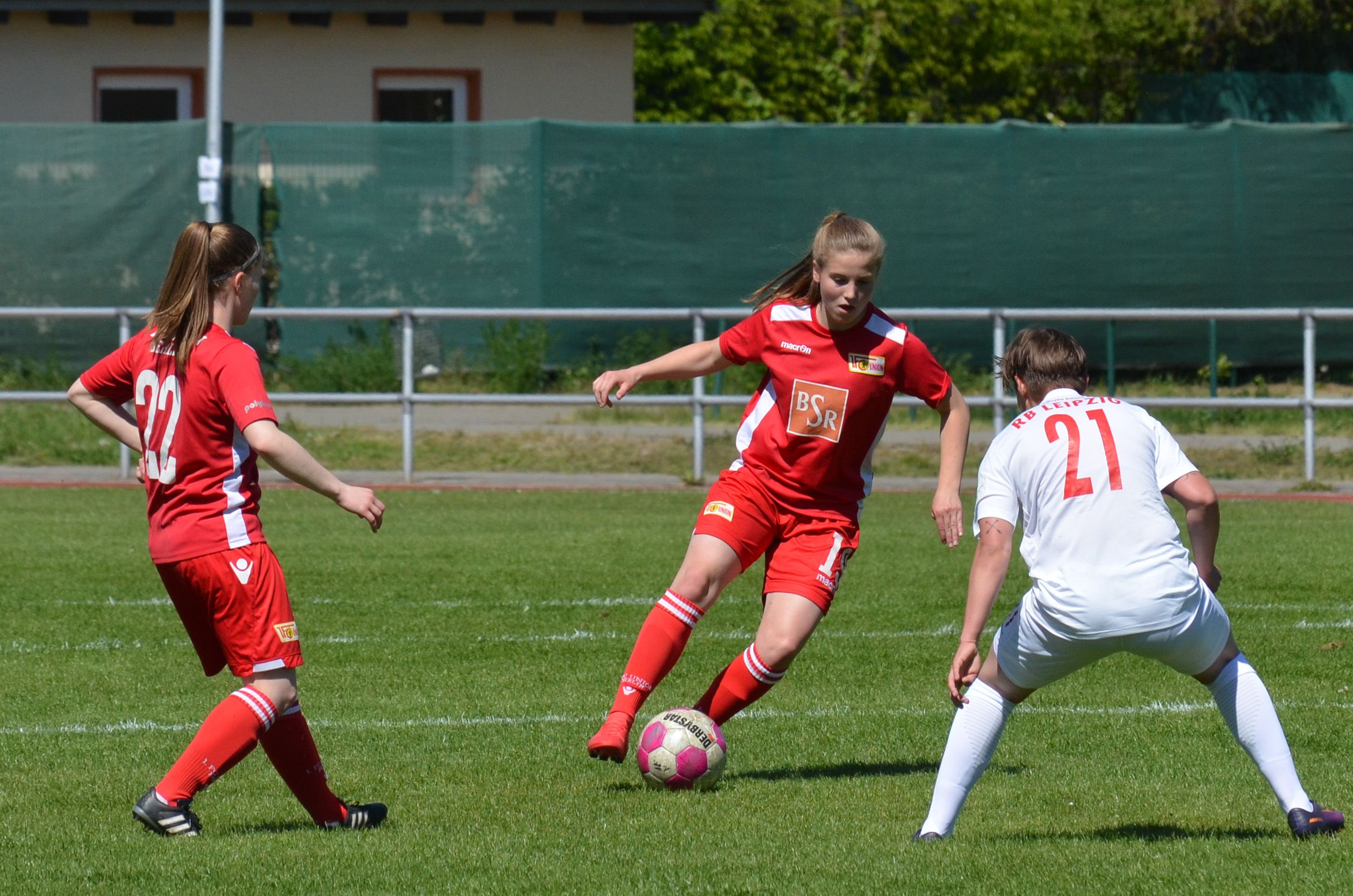 Zwei Spielerinnen tragen rote Trikots. Ein Spieler trägt Weiß. Er will den Ball bekommen. Sie spielen Fußball.
