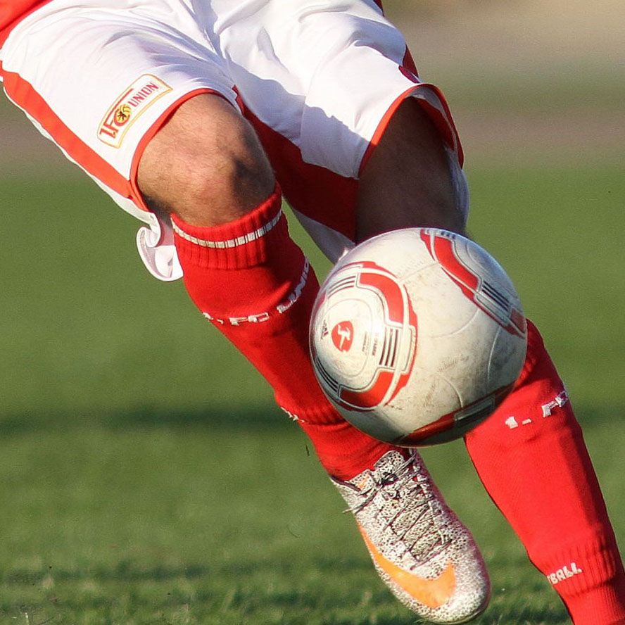 Ein Fußballspieler in roten Shorts und weißen Socken dribbelt einen Fußball auf einem grünen Feld.