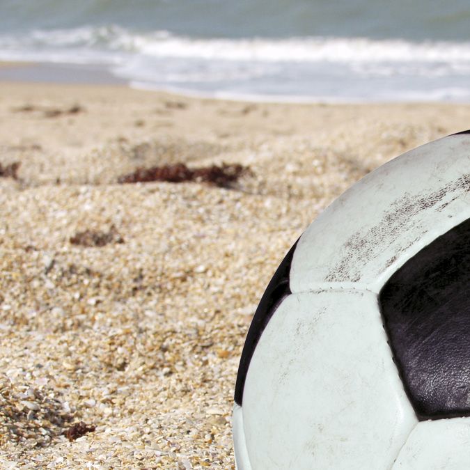 Ein Fußball liegt im Sand am Strand, mit Meer im Hintergrund.