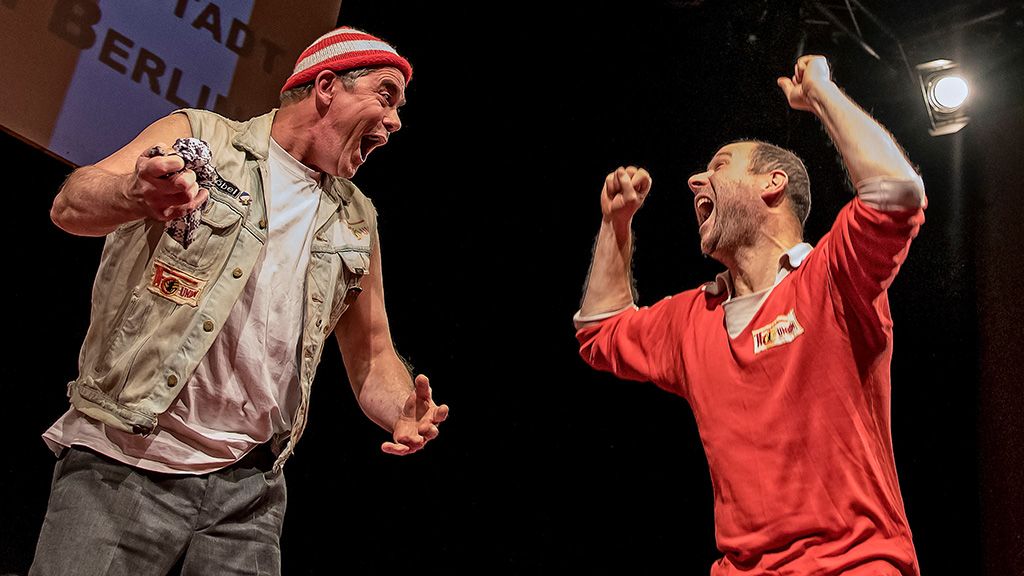 Zwei Männer auf einer Bühne, lachen und gestikulieren begeistert. Eine lebhafte Szene aus einer Performance.