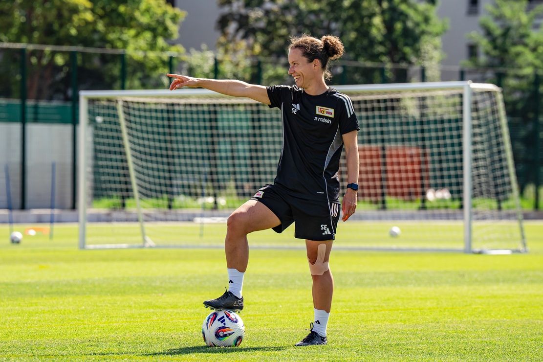 Eine Person in Sportkleidung steht auf einem Fußball und zeigt mit dem Finger, während sie auf einem Trainingsplatz steht.