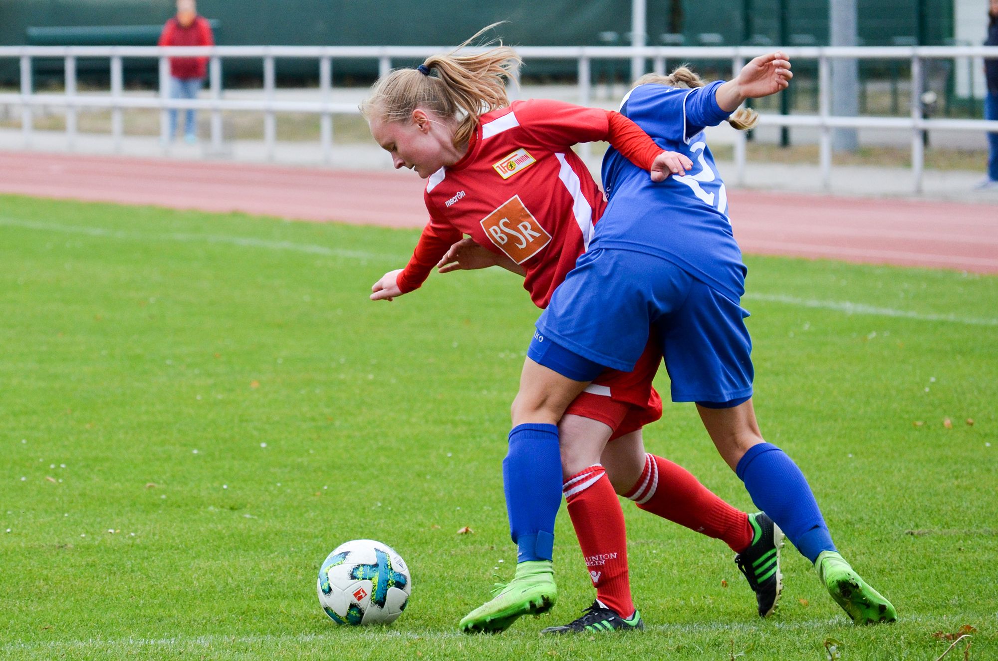Zwei Fußballspielerinnen spielen auf einem grünen Platz. Sie kämpfen um den Ball.