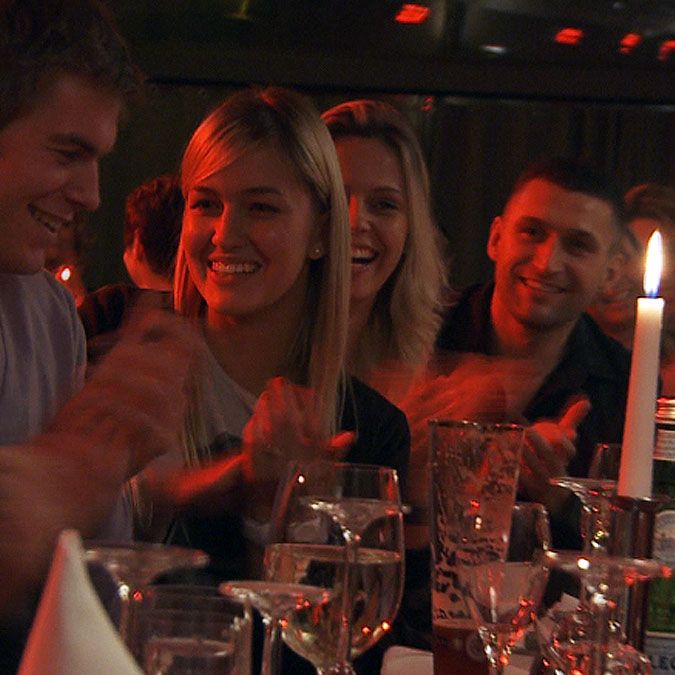 Eine Gruppe von Personen sitzt an einem Tisch, lacht und applaudiere, umgeben von Kerzenlicht und Gläsern.