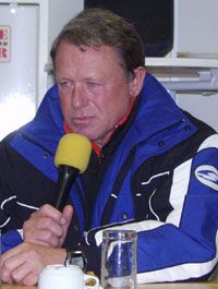 Mann mit mikrofon in blauer Jacke, der ernst schaut, sitzt an einem Tisch mit Gläsern und einem Kaffeebecher.