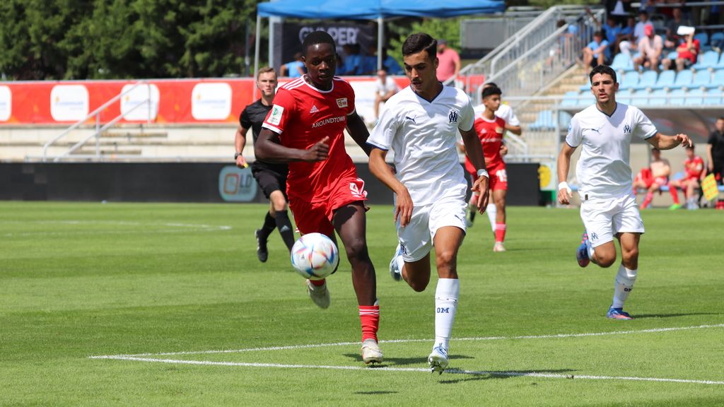 Two soccer players sprint on the field while a third player watches in the background.