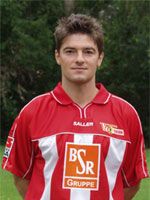 Junger Mann in rotem Fußballtrikot mit weißen Streifen, steht vor einer grünen Kulisse.