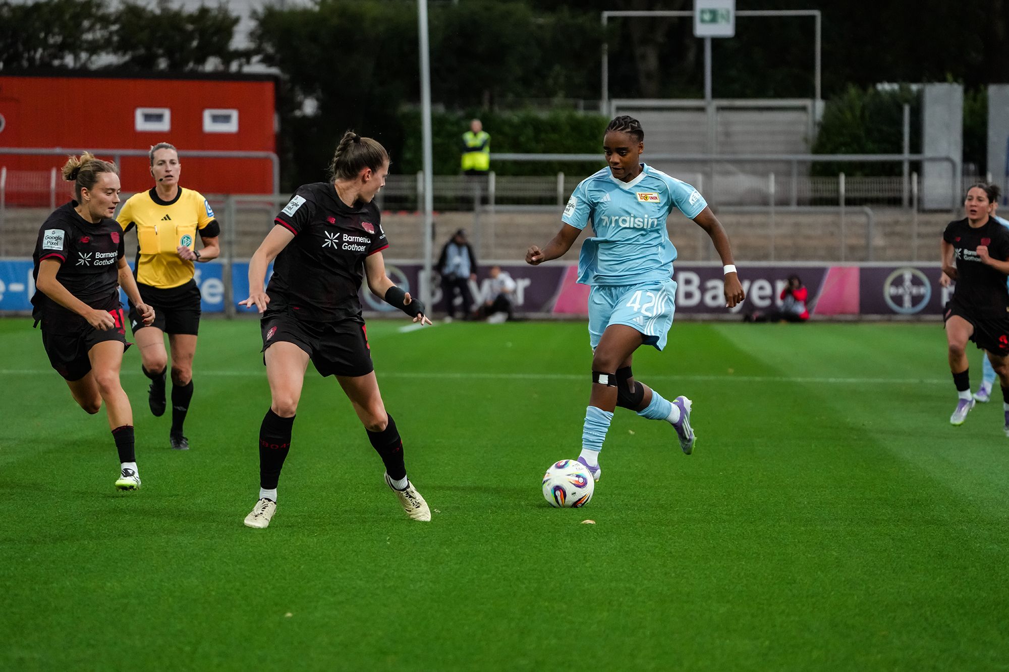 Zwei Fußballspielerinnen im Wettkampf um den Ball auf einem Rasenplatz, während eine Schiedsrichterin zuschaut.