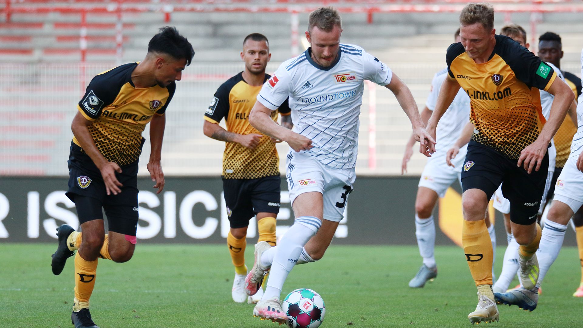 Game scene in football with a player in a white shirt being attacked by two opposing players in yellow and black.
