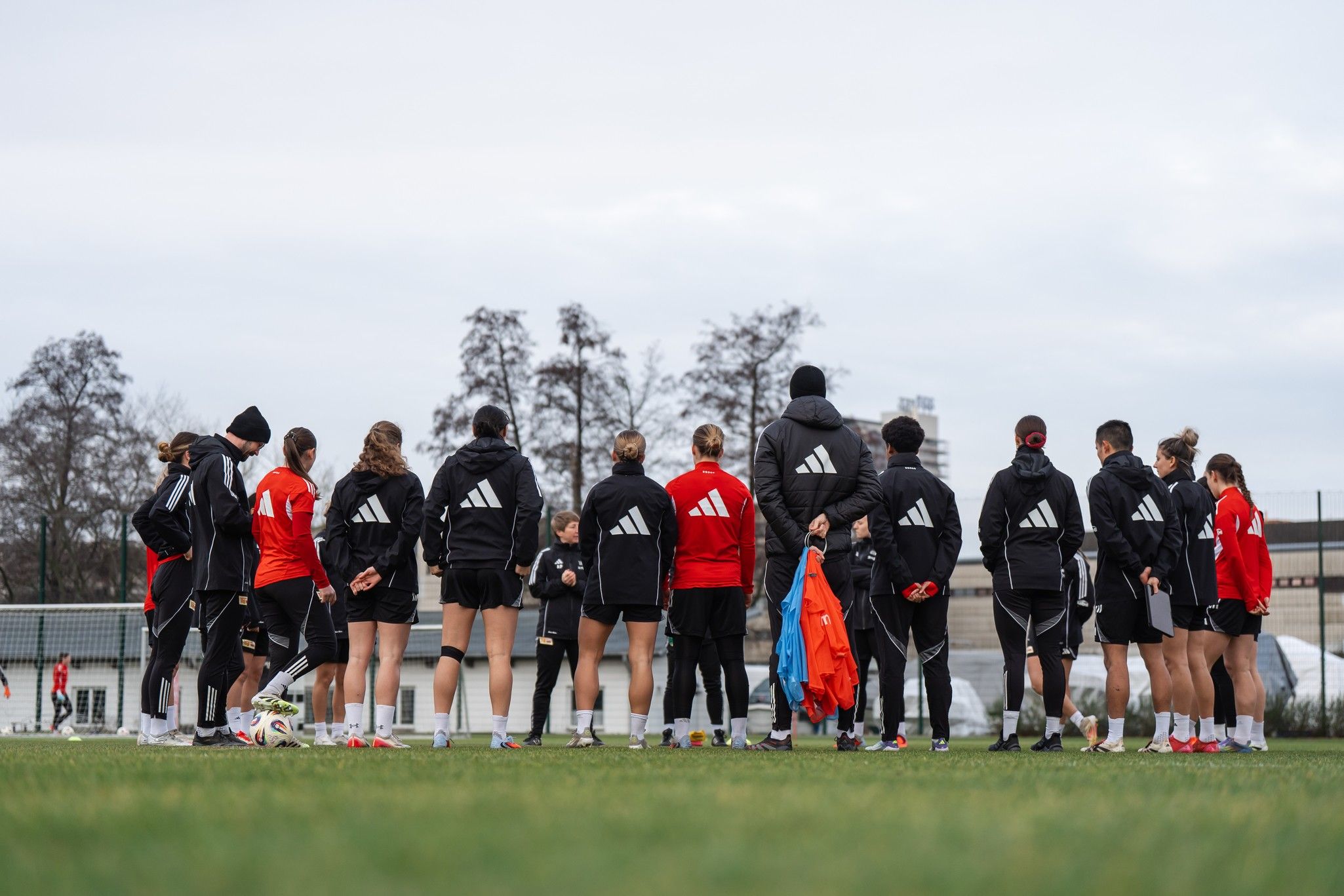 Eine Gruppe von Fußballspielern in Trainingskleidung versammelt sich auf dem Spielfeld. Im Hintergrund sind Bäume und ein Tor zu sehen.