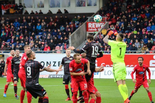 Football scene with players in red and black jerseys, goalkeeper catching the ball during a shot. Spectators in the background.