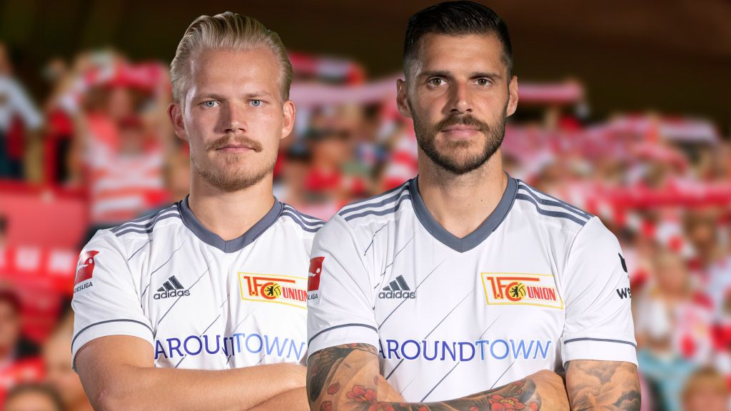 Zwei Fußballspieler tragen weiße Trikots. Sie stehen nebeneinander. Im Hintergrund jubelt die Menge.