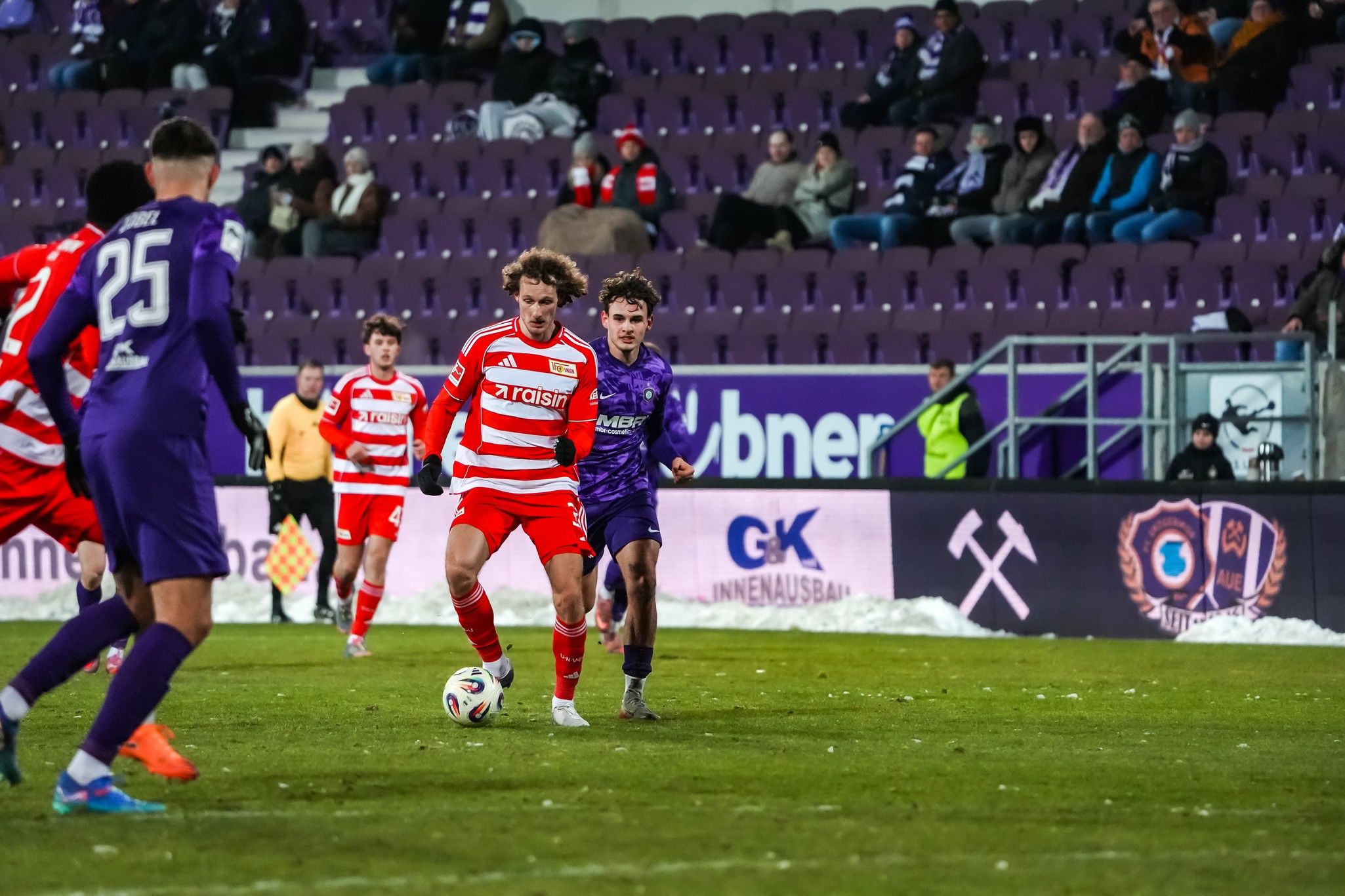 Ein Fußballspiel mit Spielern in roten und lila Trikots auf einem Rasenplatz. Zuschauer sitzen im Hintergrund in einem Stadion.