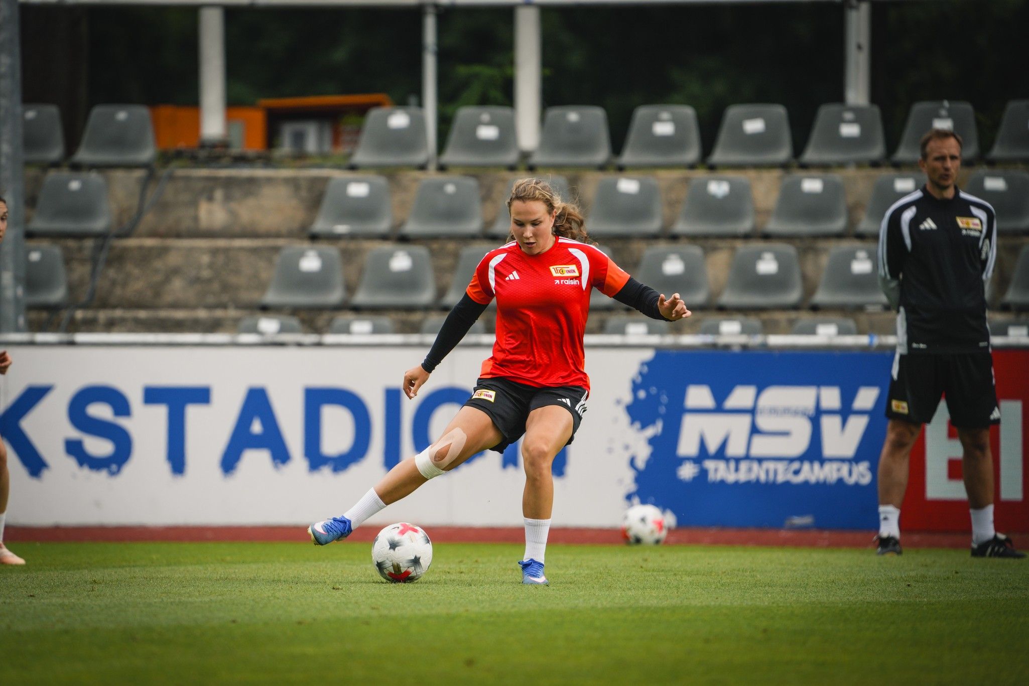 Eine Spielerin in rotem Trikot tritt einen Fußball auf einem Trainingsplatz, während andere Spieler im Hintergrund zuschauen.