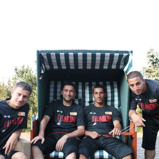 Vier Männer in einheitlichen schwarzen T-Shirts sitzen lächelnd in einem Liegestuhl im Freien.