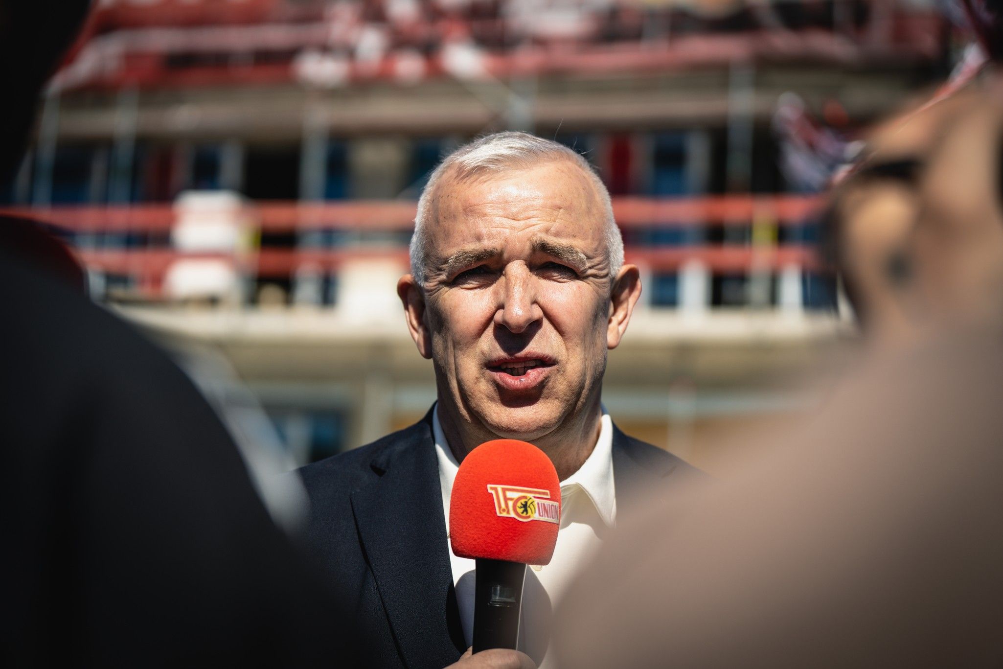 Mann mit Mikrofon und rotem Logo spricht vor einem Baustellen-Hintergrund.