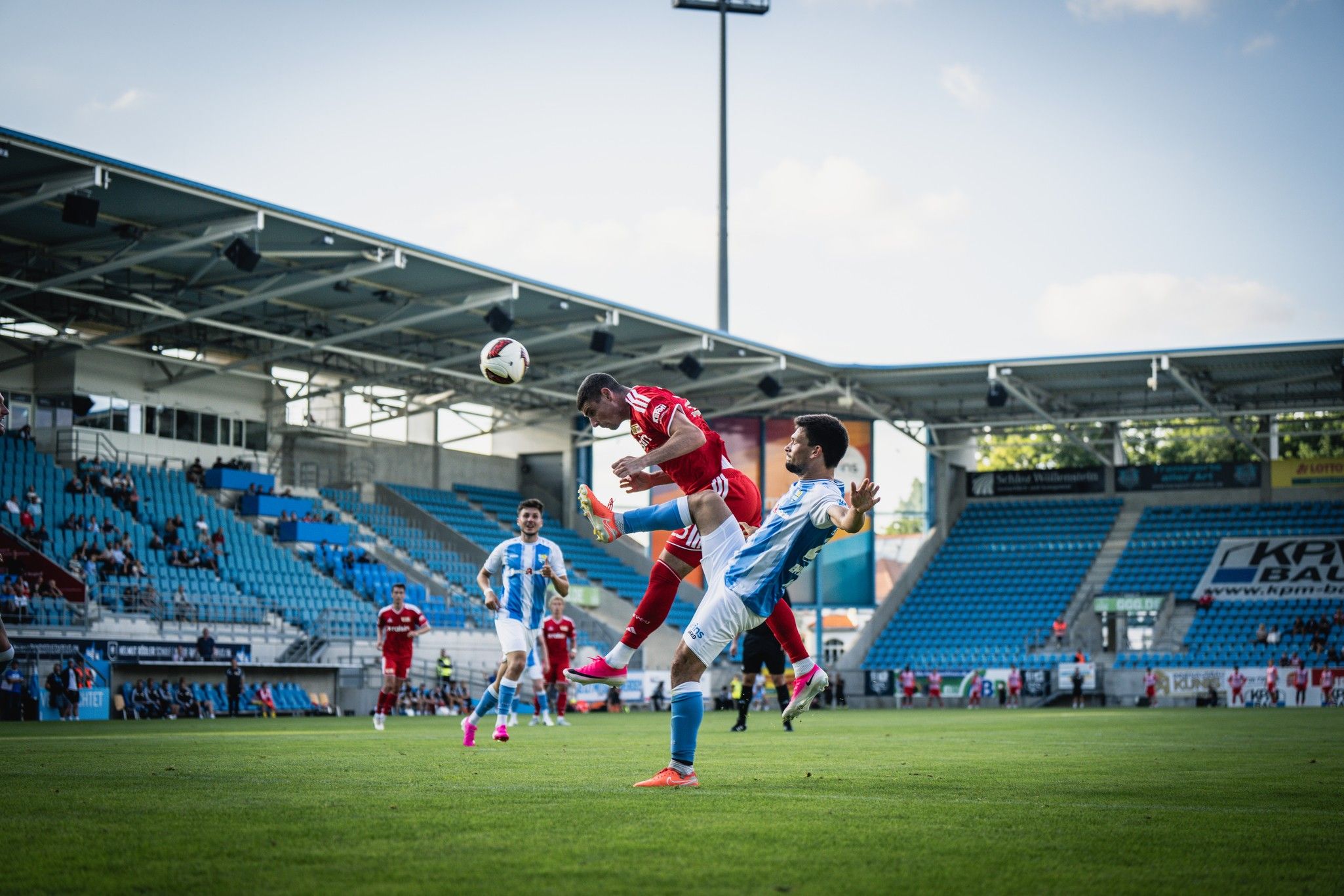 Zwei Fußballspieler im Sprung, der eine versucht, den Ball zu köpfen, während der andere ihn am Bein berührt. Stadionhintergrund mit Zuschauern.