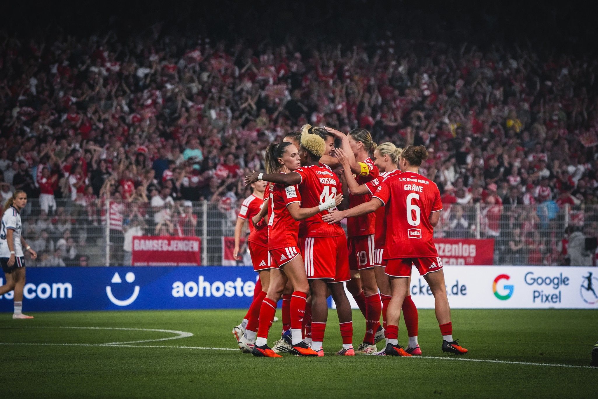 Spielerinnen eines Fußballteams feiern ein Tor in einem Stadion mit jubelndem Publikum im Hintergrund.