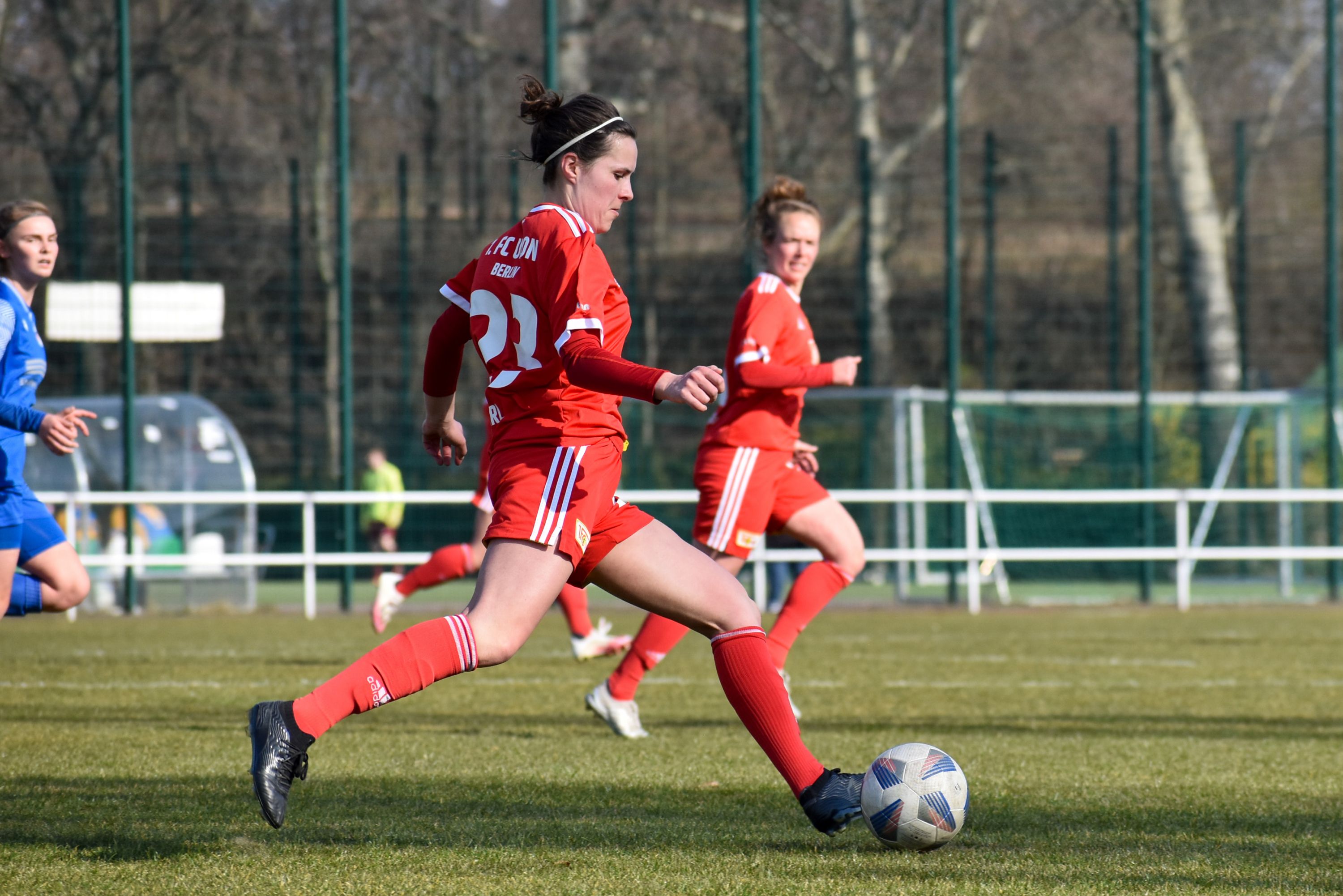 Eine Spielerin in rotem Trikot läuft mit dem Ball auf einem Fußballfeld. Im Hintergrund sind weitere Spielerinnen zu sehen.