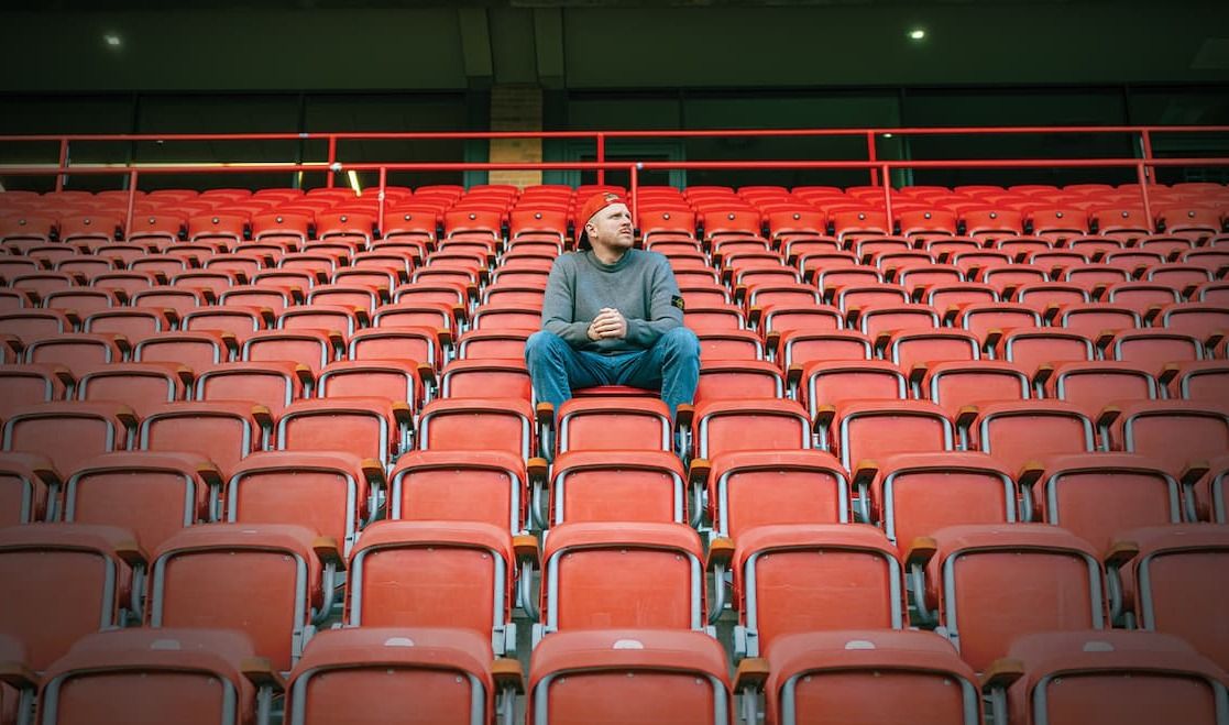 Ein Mann sitzt allein auf einer leeren Tribüne mit roten Sitzen im Stadion.