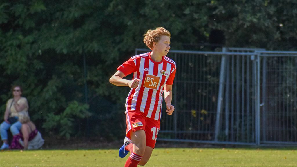 Ein junger Fußballspieler in rot-gestreifter Uniform läuft auf dem Spielfeld. Hintergrund mit Bäumen und Zuschauern.