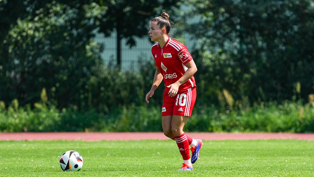 A woman in a red football shirt stands on a field preparing for a pass or a shot.