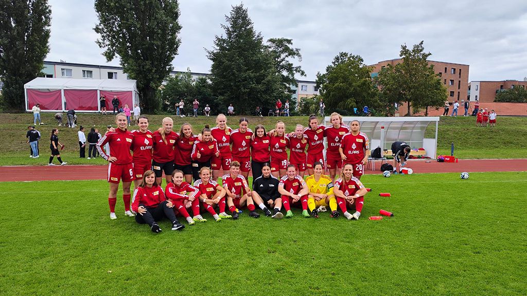 Frauenfußballmannschaft in roten Trikots posiert im Freien auf einem Rasenfeld, Zuschauer im Hintergrund.