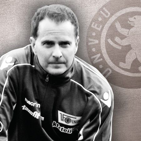 Schwarz-weißes Porträt eines Mannes in Sportkleidung, hinter ihm ein Logo eines Fußballvereins.