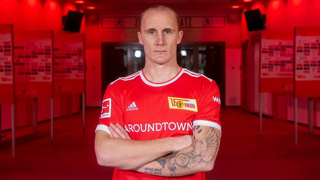 Ein Fußballspieler in rotem Trikot mit Tattoos steht selbstbewusst in einem rot beleuchteten Raum.