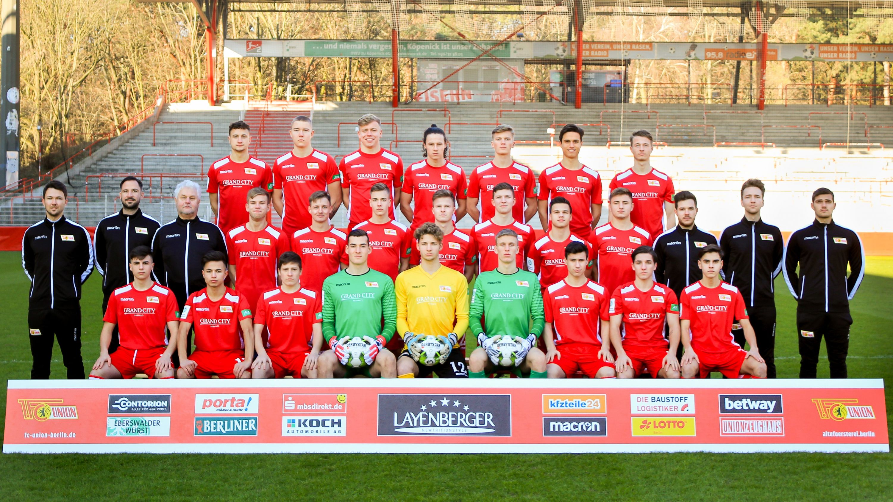 Mannschaftsbild einer Fußballmannschaft in roten Trikots, gruppiert auf einem Rasenplatz vor den Tribünen.