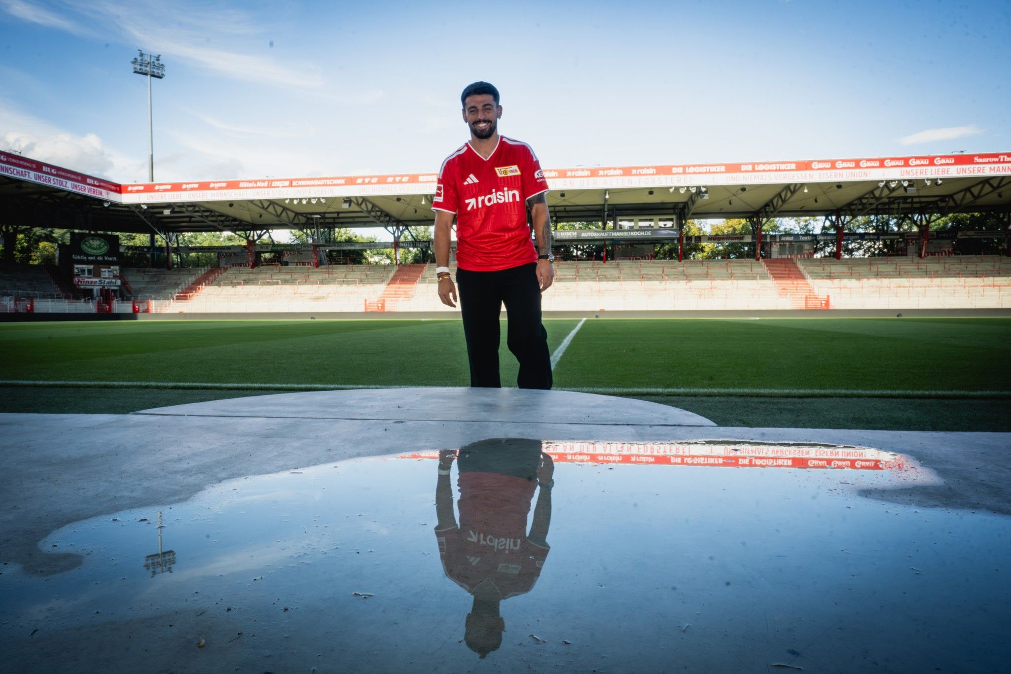 Ein Fußballspieler in rotem Trikot steht auf einem Spielfeld, der Himmel spiegelt sich in einer Pfütze vor ihm.