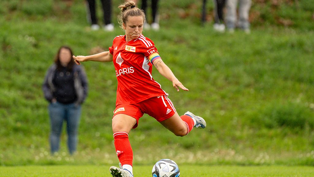 A player in a red soccer jersey kicks a ball on the field, with spectators on the grass in the background.