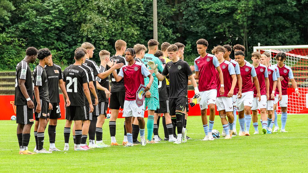 Zwei Gruppen von Fußballspielern in Trikots schütteln sich die Hände auf einem Spielfeld vor einem Tor.