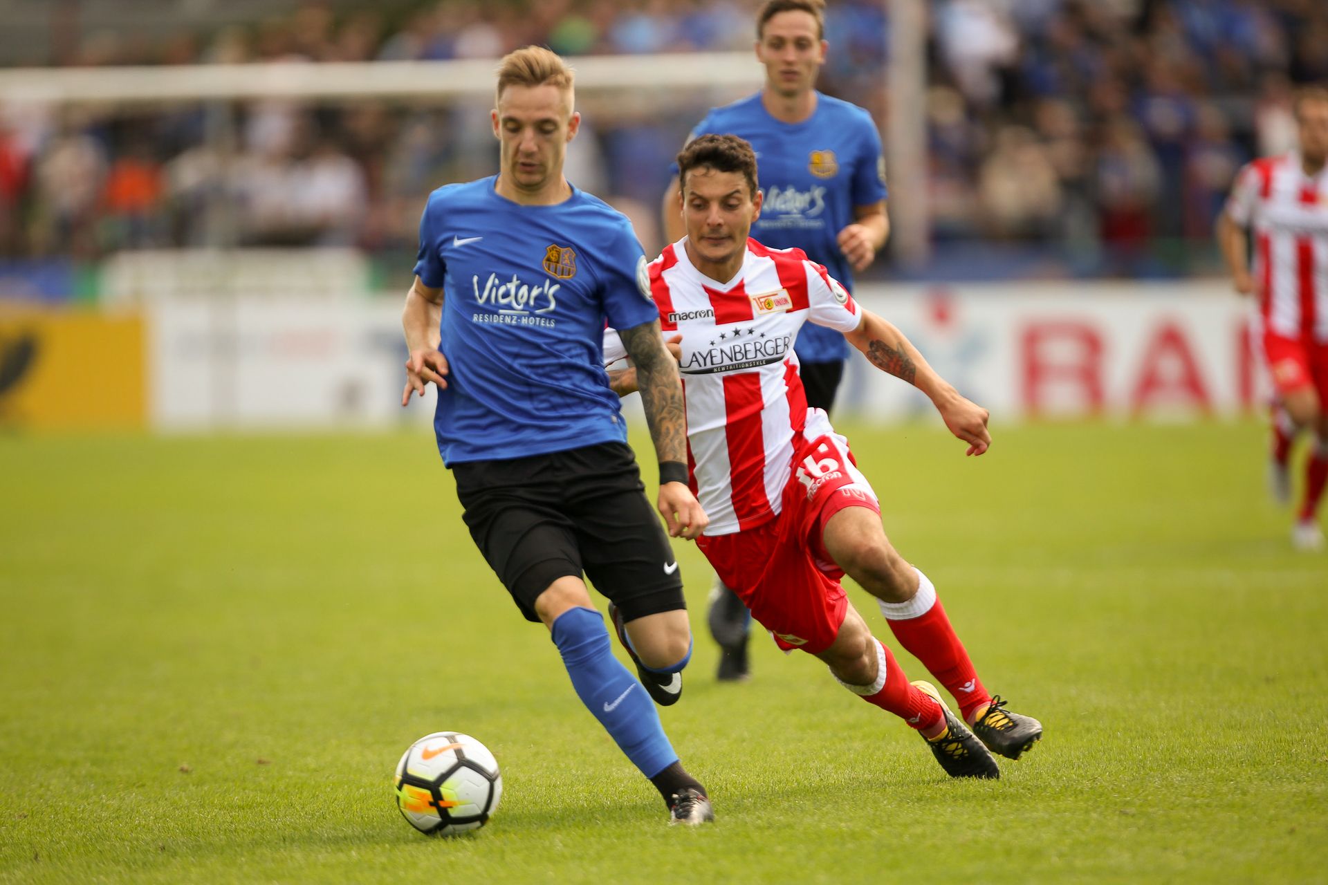Zwei Fußballspieler rennen um den Ball. Der eine trägt Blau. Der andere hat rot-weiß gestreifte Kleidung. Sie sind auf einem grünen Feld.