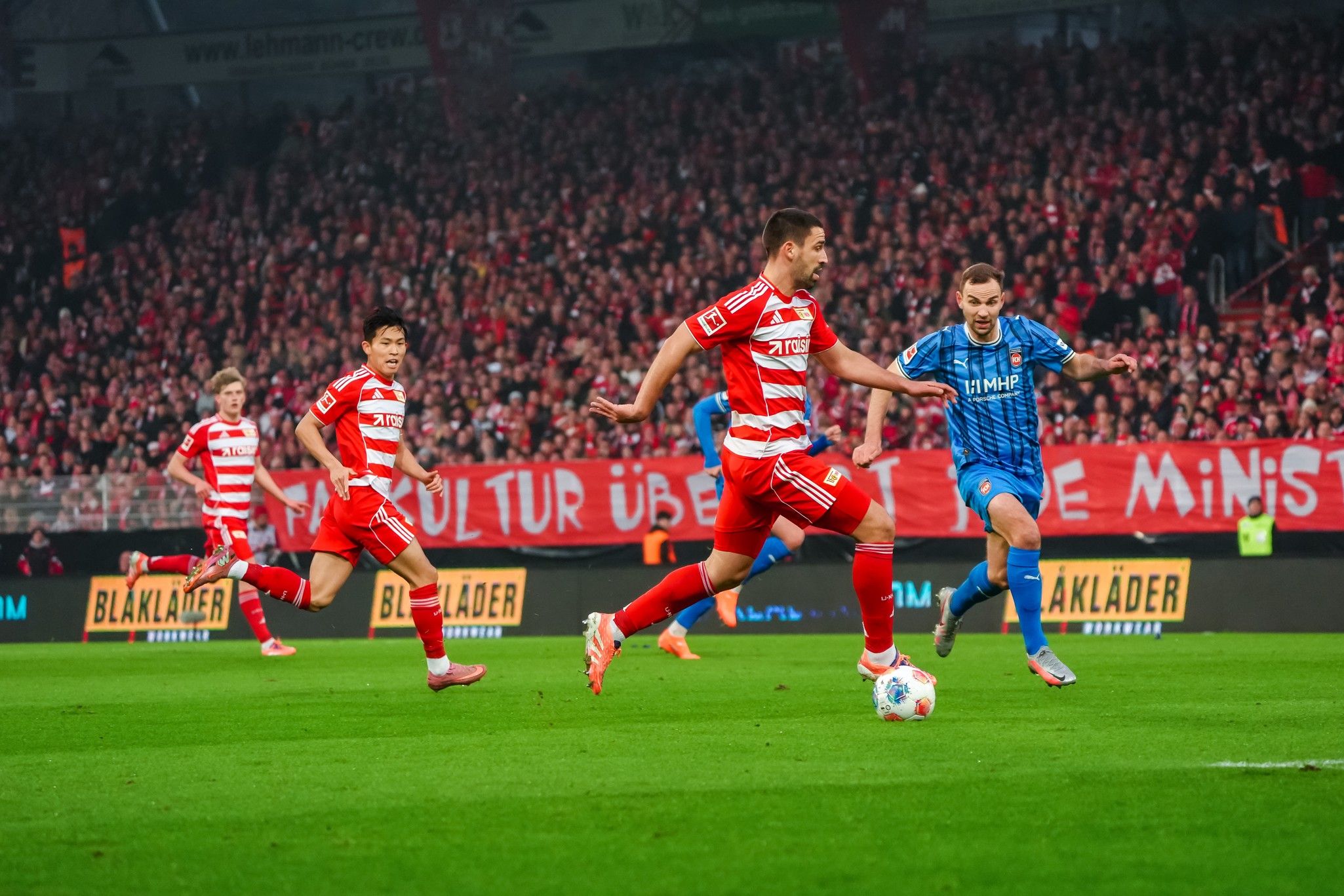 Spielszene eines Fußballspiels mit drei Spielern in roten und blauen Trikots auf einem Rasenplatz vor einer begeisterten Zuschauermenge.