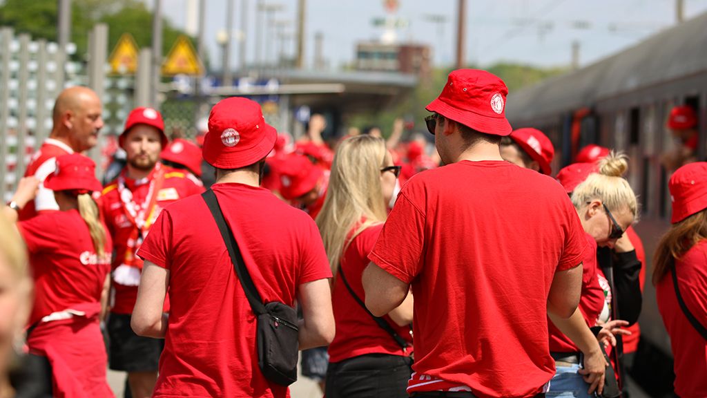 Menschen in roten T-Shirts und Hüten versammeln sich an einem Bahnhof, umgeben von Verkehrsschildern und einem Zug.