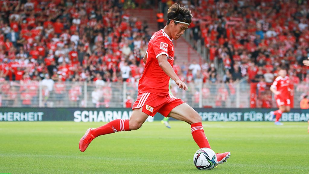 Spieler in rotem Trikot schießt den Ball auf einem Fußballfeld, umgeben von jubelnden Fans in der Menge.