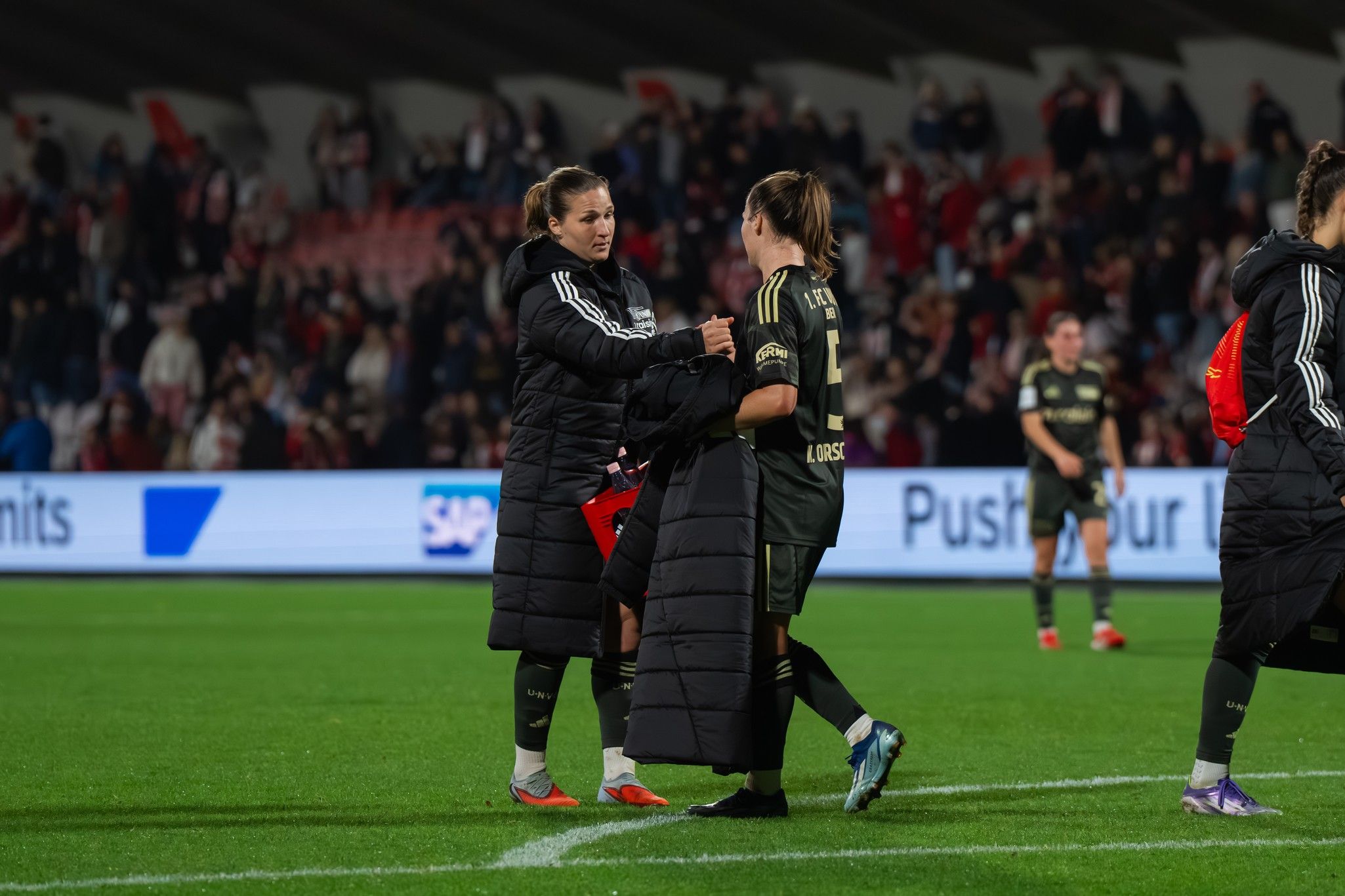 Zwei Fußballspielerinnen in schwarzen Jacken stehen auf dem Spielfeld, umgeben von jubelnden Fans im Hintergrund.