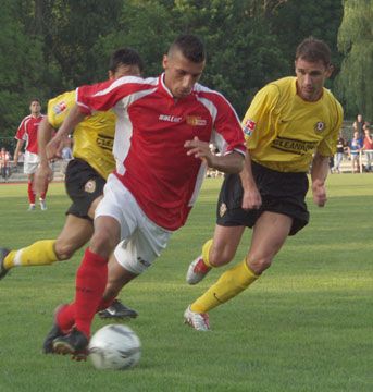 Ein Fußballspieler in rotem Trikot dribbelt den Ball, während zwei Spieler in gelben Trikots ihn verfolgen.