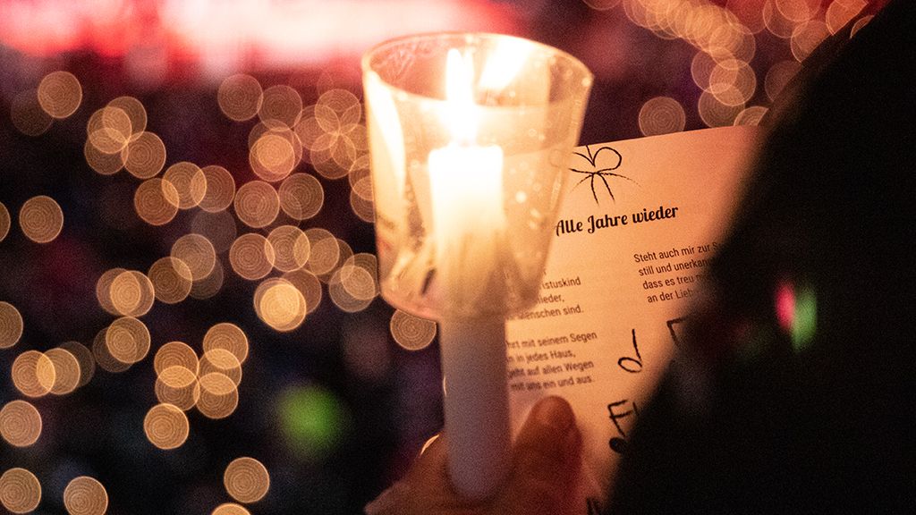Eine Hand hält eine Kerze vor einem verschwommenen Hintergrund von Lichtern, während ein Liedtext in der anderen Hand sichtbar ist.